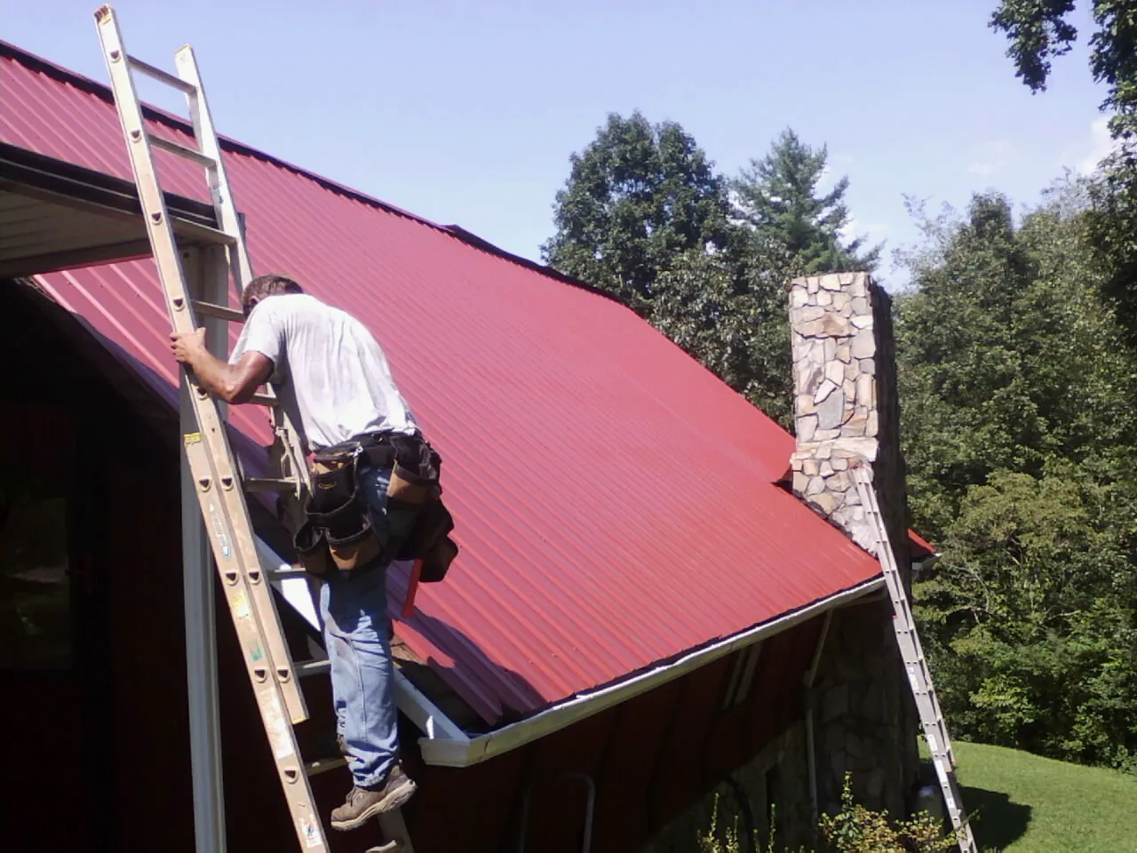 Roofer on a ladder beside a red metal roof and stone chimney — Soffit & Fascia Repair by Roofing Brothers in Mound